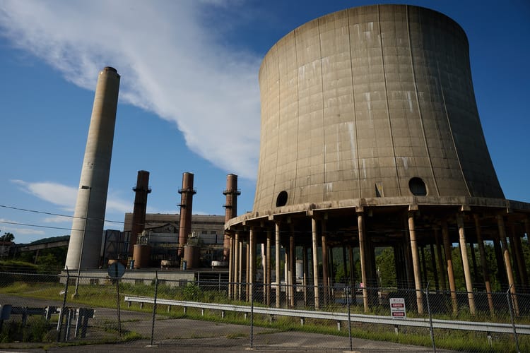 A hulking cooling tower in worn concrete looms in front of a larger plant building with smokestacks
